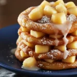 Baked apple fritters stacked on a blue plate, topped with diced apples, powdered sugar, and a light vanilla glaze dripping over the edges.