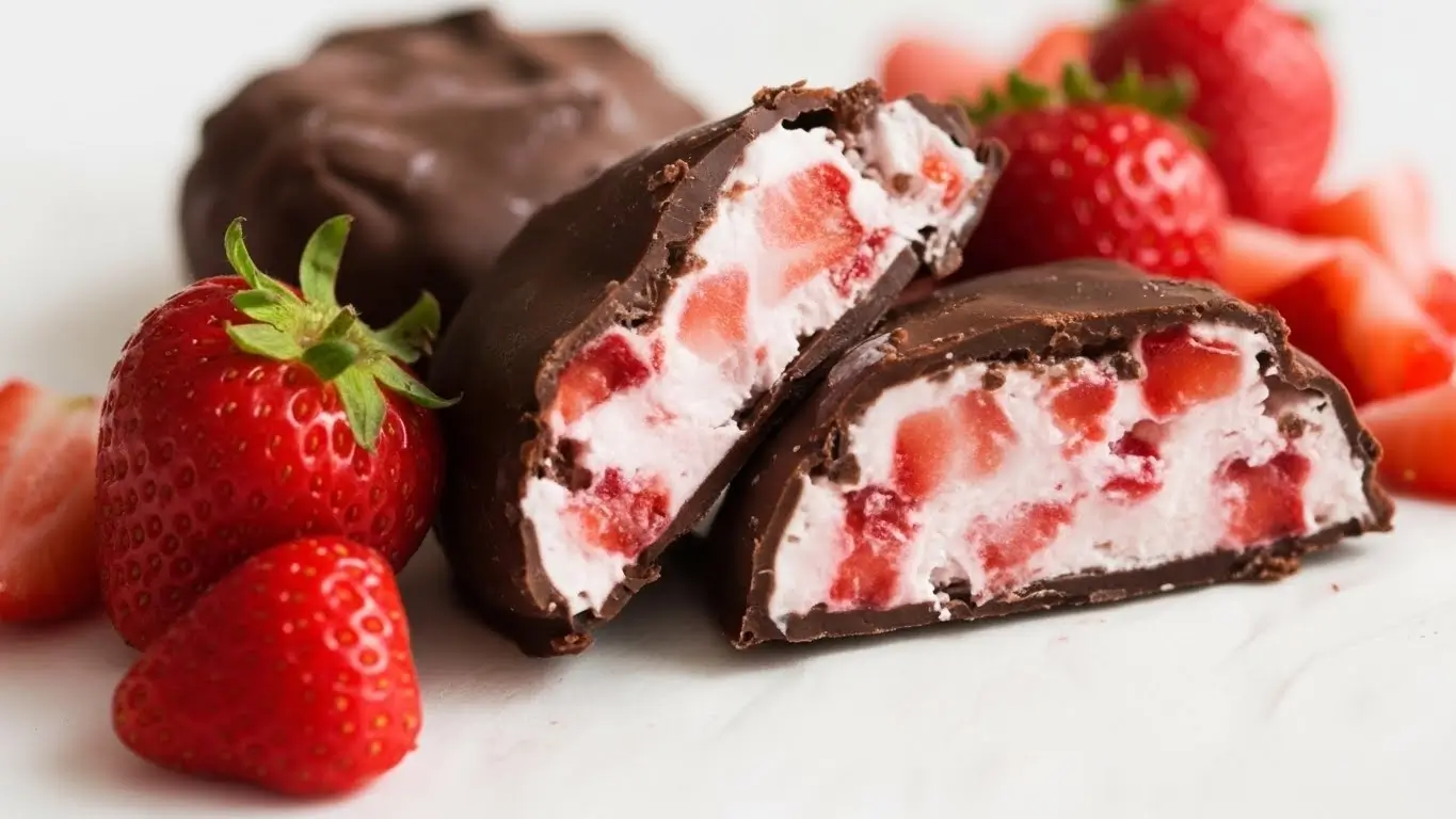 Chocolate strawberry yogurt clusters arranged on a white plate atop a clean white countertop, coated in smooth chocolate with visible strawberry pieces.